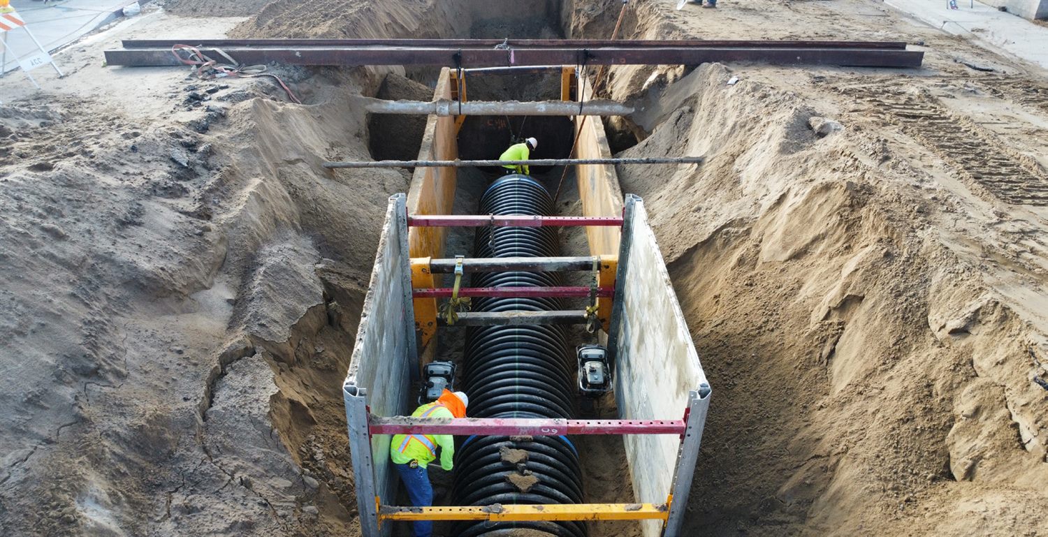 Crews installing a large storm drain pipe in an excavated trench